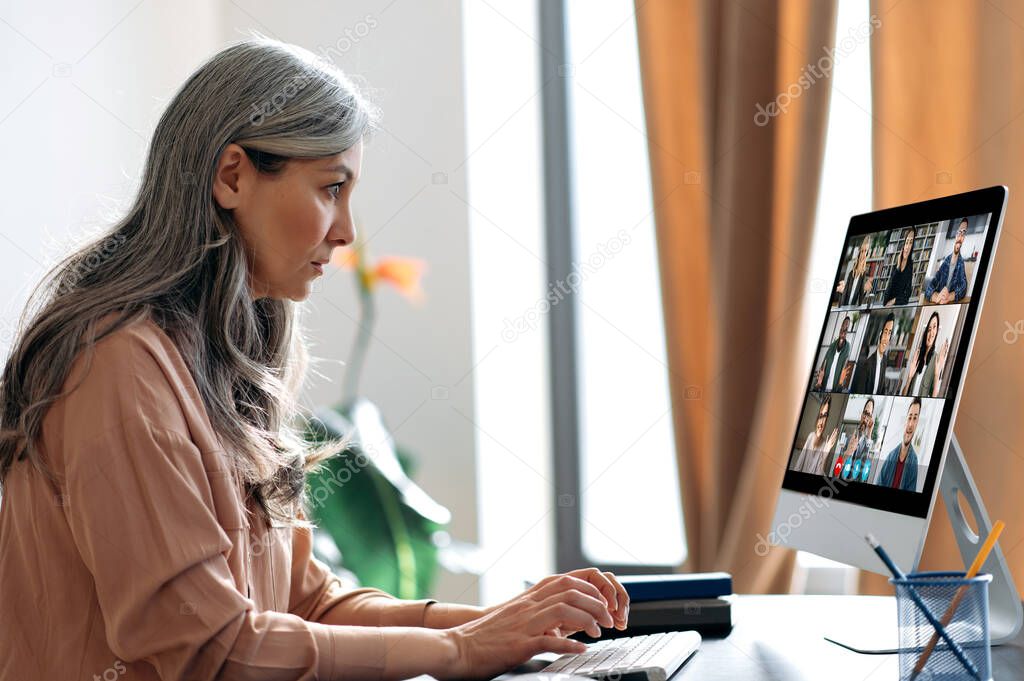 Virtual meeting, online business meeting. Serious asian mature gray-haired woman communicate with business partners by video call uses app and computer, sitting at her work desk