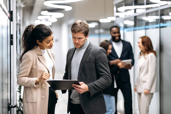 Serious multiracial colleagues standing in office hall, talking about work questions. Concentrated business partners discussing corporate project, giving each other advices, teamwork concept