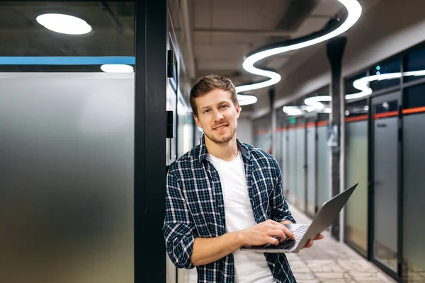 Portrait of attractive male freelancer in stylish wear at the modern office. Caucasian young businessman holds laptop in arms, looks at the camera, smiling, working in a creative office