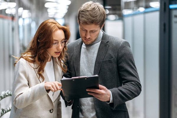 Focused young adult coworkers discussing new business plan, standing in office hall, working together. Handsome male employee ask an advice from beautiful redhead woman colleague, partnership concept