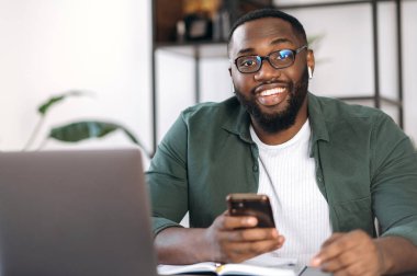 Dost canlısı, gözlüklü, çalışma masasında oturan, elinde akıllı bir telefonla kameraya gülümseyerek bakan Amerikalı bir gencin portresi.