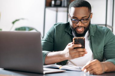 Kendine güvenen Afro-Amerikan bir adam, masasında oturuyor, çalışırken cep telefonu kullanıyor, internette geziniyor, e-postalara cevap veriyor, proje fikirleri arıyor.