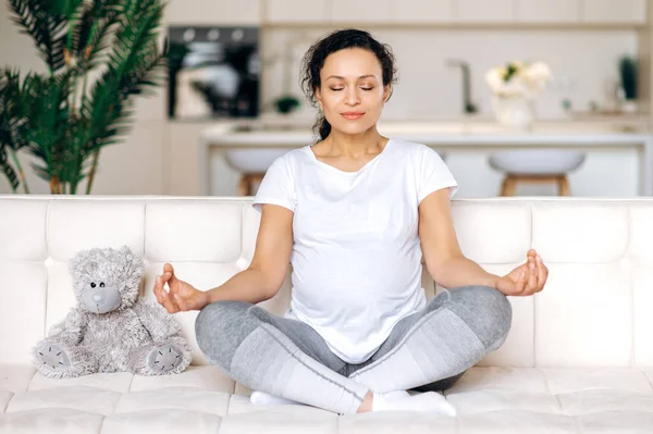 Attractive contented, beautiful future mom, brunette, mixed race, sitting on sofa at home in living room, in lotus position with closed eyes relaxes and meditates, focusing on exercises