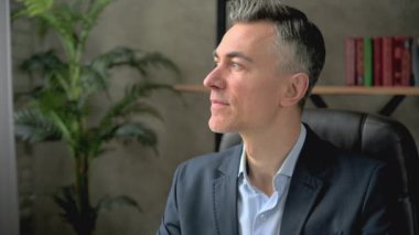 Resmi takım elbiseli yakışıklı, beyaz saçlı, beyaz saçlı, iş adamı ya da avukatın yakın plan portresi, çalışma masasında oturuyor, önce yana bakıyor, sonra kameraya arkadaşça gülümsüyor.