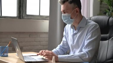 Sağlıkla, önlemlerle ilgilenir. Kafkasyalı bir adam, ofis çalışanı ya da yöneticisi, tıbbi bir maske takıyor, resmi kıyafetlerle iş yerinde oturuyor, iş sırasında ellerini antiseptikle dezenfekte ediyor, prezervatif kullanıyor.