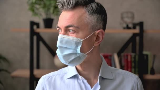 Βίντεο πορτραίτο ενός μεσήλικα καυκάσιου γκριζομάλλη άνδρα, υπάλληλος γραφείου, διευθυντής, επιχειρηματίας, φορώντας μια ιατρική προστατευτική μάσκα κατά τη διάρκεια της καραντίνας, κοιτάζει την κάμερα. Έννοια υγειονομικής περίθαλψης — Αρχείο Βίντεο