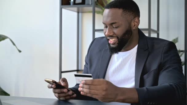 満足笑顔アフリカ系アメリカ人の男は、スーツで、自宅で作業机に座って、携帯電話とクレジットカードを手に入れ、オンライン購入のために支払い、カード番号を入力してくださいオンラインショッピング、注文配達 — ストック動画