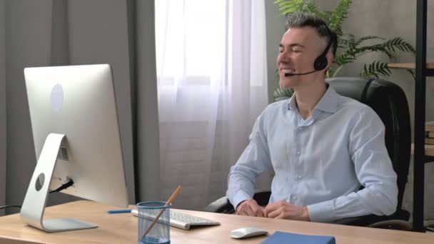 Mutlu Kafkasyalı yetişkin erkek, çalışan, yönetici ya da CEO, resmi gömlek ve kulaklıkla ofiste oturuyor, online bir iş toplantısı için bilgisayar kullanıyor, iş arkadaşlarıyla iletişim kuruyor, gülümsüyor. — Stok video