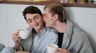 Homoseksüel ilişki. Birbirine aşık iki eşcinsel birlikte vakit geçiriyorlar, birbirlerine nazikçe sarılıyorlar ve birbirlerini okşuyorlar, mutfakta yerde oturmuş, kahve fincanlarıyla üzerlerini ekoseyle örtüyorlar.