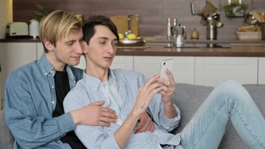 Aşık mutlu eşcinsel çift evde birlikte vakit geçiriyor. İki homoseksüel adam oturma odasında kanepede uzanıp akıllı telefon kullanıyor, internette geziniyor, sosyal medyada sohbet ediyor, online alışveriş yapıyor, gülümsüyor.