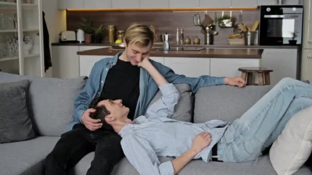 Iubesc cuplul gay acasă. Băieții petrec timp împreună acasă pe canapea, unul stă, celălalt se află în poala lui, se îmbrățișează și se mângâie reciproc, sunt fericiți să fie împreună, zâmbind — Videoclip de stoc