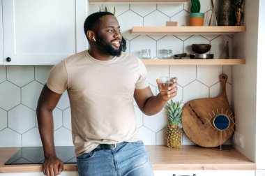 Mutlu, sağlıklı, Afro-Amerikan sakallı adam, mutfakta duruyor, günlük kıyafetlerle, elinde bir bardak temiz suyla, yan tarafa bakıyor, arkadaşça gülümsüyor. Sağlıklı yaşam tarzı kavramı