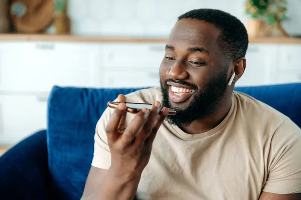 Close-up of handsome friendly confident african american guy sitting in living room on sofa talking or recording audio message on smartphone for friend or colleague, smiling happily