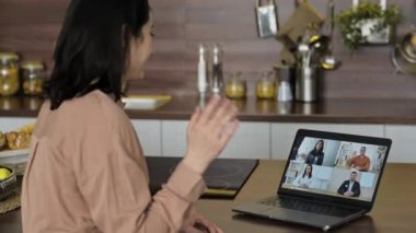 Görüntülü konuşma, çevrimiçi iletişim. Serbest çalışan ya da öğrenci bir kız mutfakta oturuyor, video konferansında iletişim kurmak için dizüstü bilgisayar kullanıyor, interlocutorları selamlıyor, çalışıyor ya da evden uzaktan çalışıyor.