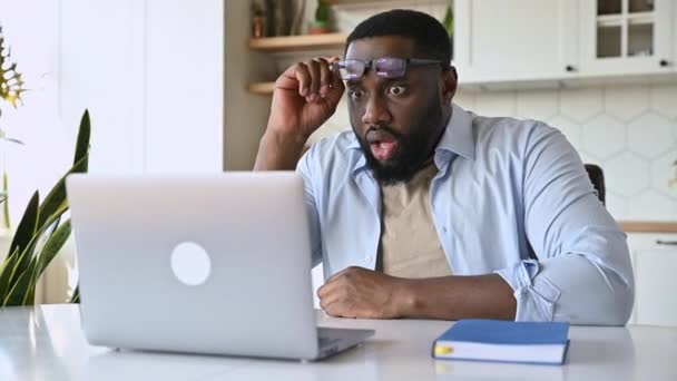 Zszokowany Afroamerykanin brodaty biznesmen lub freelancer, siedzi przy biurku, patrzy zaskoczony na swojego laptopa, czyta nieoczekiwane wiadomości, zdejmuje okulary, zdezorientowany wyraz twarzy — Wideo stockowe