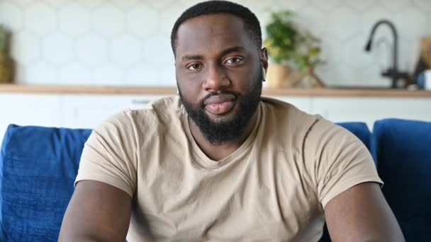 Close-up of handsome happy calm young african american bearded man, sitting on sofa at home in living room, wearing t-shirt, looking at camera and smiling friendly — Stock Video
