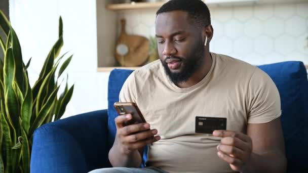 Zakupy online. Szczęśliwy Afroamerykanin, w stylowych ciuchach, siedzący na kanapie, trzymający smartfona i kartę kredytową, gestykulujący rękami, uśmiechający się szczęśliwie, dostał zniżkę na zakupy — Wideo stockowe