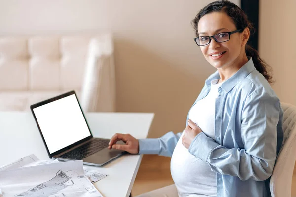 A pregnant woman, designer or freelancer, sits at a work desk, gently hugs her tummy with her hand, looks and smiles into camera, on the table a laptop with blank white screen and a working documents