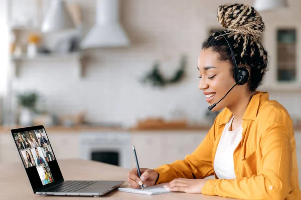 Happy African American Siswa perempuan mendengarkan kuliah online menggunakan laptop dan headset, mencatat, studi jarak jauh, guru dan siswa lainnya berada di layar laptop. E-learning — Stok Foto
