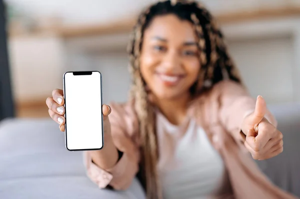 Terpencil african Amerika tersenyum wanita muda, melihat ke kamera, memegang smartphone dengan layar putih kosong di tangan, menunjukkan jempol ke atas gerakan, salinan ruang — Stok Foto