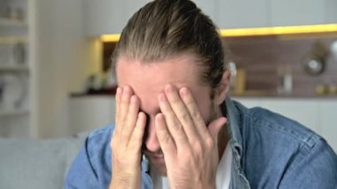 Baş ağrısı, migren. Kafkasyalı, başı ağrıyan üzgün bir adam, elleri kapalı bir şekilde şakaklarının yanında oturuyor, kanepede oturuyor, çalışmaktan sıkılmış, kişisel sorunları için endişeleniyor, stresli.