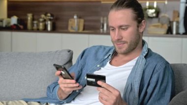Tatmin olmuş bir beyaz adam, günlük kıyafetleriyle, evinde bir kanepeye oturur, elinde bir telefon ve kredi kartı tutar, online alışverişleri veya siparişleri öder, kart numarasına girer, online alışveriş yapar,
