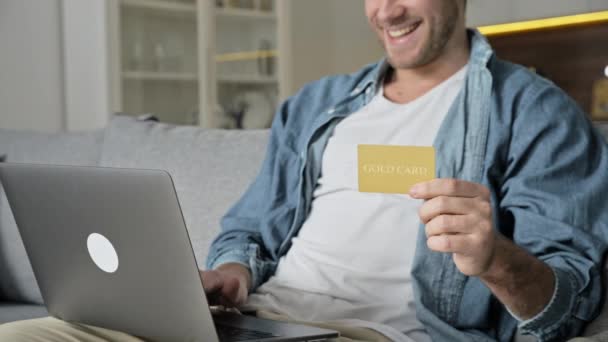 Szczęśliwy uśmiechnięty biały młodzieniec w luźnym ubraniu, używa laptopa i karty kredytowej do zakupów online, siedząc w domu na kanapie, płaci za zakupy online lub dostawę towarów, wpisuje dane karty — Wideo stockowe
