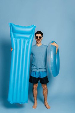 Mavi şişme yüzüklü ve şilteli, güneş gözlüklü, izole edilmiş mavi bir arka planda duran beyaz tişörtlü adam kameraya bakar ve gülümser ve tatil konseptine.