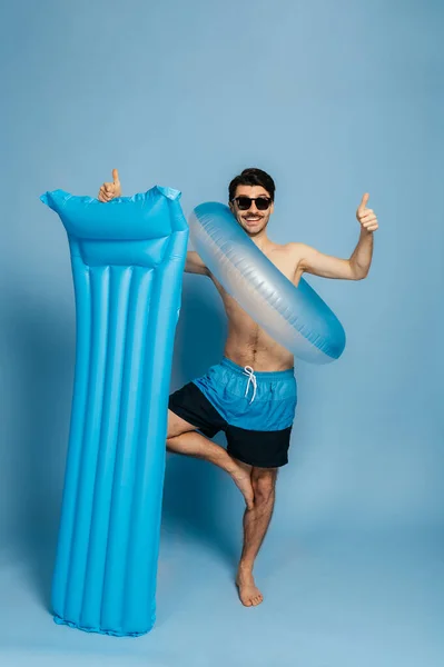 Teljes hosszúságú boldog kaukázusi vonzó ing nélküli férfi turista standok kék felfújható gyűrű és matrac egy elszigetelt kék háttér napszemüveg, mutatja hüvelykujj fel és mosolygós, nyaralás koncepció — Stock Fotó
