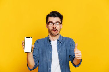 Aptal yüz ifadesi olan komik giyimli bir adam sunumunuz için beyaz ekranlı akıllı telefonu tutuyor, başparmağını kaldırıyor, izole edilmiş turuncu arka planda duruyor.