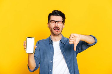 Beyaz tenli, yüzünde negatif ifade olan beyaz bir adam sunumunuz için beyaz ekranlı akıllı telefonu tutuyor, baş parmağını aşağı gösteriyor, izole edilmiş turuncu arka planda duruyor, negatif duygu.