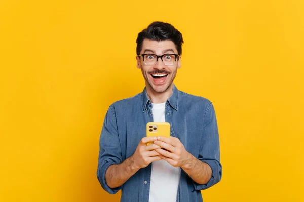 Omráčený stylový chlap s brýlemi, pomocí smartphonu, chatování online, psaní vzkazů, prohlížení internetu, sociálních médií, vypadá překvapeně na kameru, stojí na izolovaném oranžovém pozadí — Stock fotografie