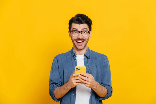 Šťastný pohledný úžasný stylový chlap s brýlemi, pomocí smartphonu, chatování online, psaní vzkazu, prohlížení internetu, sociálních médií, vypadá překvapeně na kameru, stojí na izolované oranžové — Stock fotografie