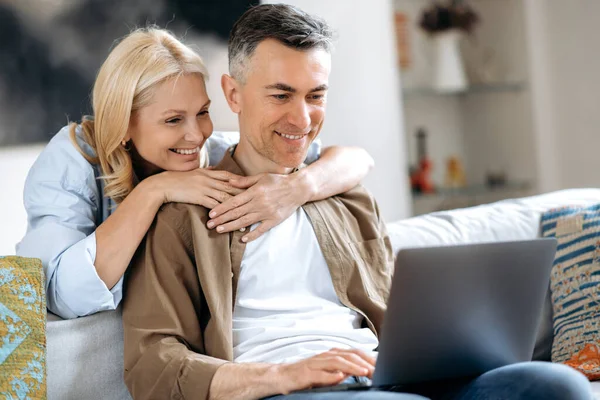 A middle aged married happy caucasian couple at home on the couch is using laptop. Joyful husband and wife browsing internet, social media, chatting with friends, do online shopping for home, smiling