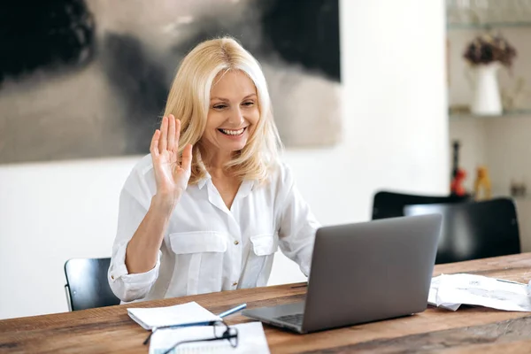 Blanke aantrekkelijke elegante volwassen zakenvrouw in een stijlvol wit shirt zittend op een werkplek, maakt gebruik van laptop om te communiceren met collega 's via videoconferentie, begroet gesprekspartner, vriendelijke glimlach — Stockfoto