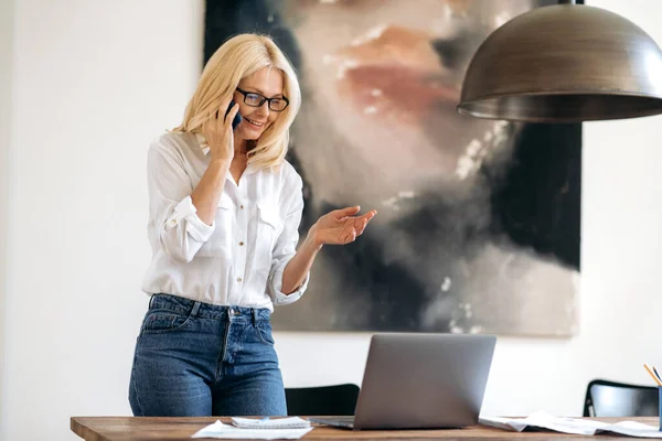 Succesvolle elegante drukke blanke zakenvrouw op het werk, stijlvol geklede volwassen blonde dame die met een collega op de smartphone staat te praten, kijkt naar laptop, glimlachend vriendelijk — Stockfoto