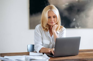 Başarılı, olgun beyaz sarışın, iş kadını, menajer ya da serbest çalışan, beyaz gömlek giyen, iş fikrinin üzerinde dizüstü bilgisayarla çalışan, strateji geliştiren, dizüstü bilgisayar ekranına bakan,