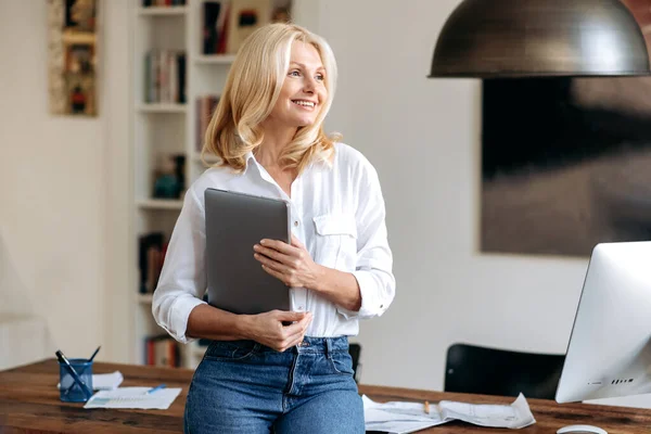Portret van een charmante, elegante blanke blonde vrouw, in een kantoor in de buurt van haar werkplek, gekleed in stijlvolle kleren, met een laptop in haar hand, naar de zijkant kijkend en lachend vriendelijk — Stockfoto