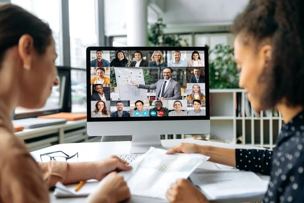 Reunião de negócios online, brainstorm. Veja sobre os ombros de duas mulheres para uma tela de computador com diferentes pessoas multirraciais bem sucedidas reunidas em uma videoconferência para discutir questões de trabalho — Fotografia de Stock