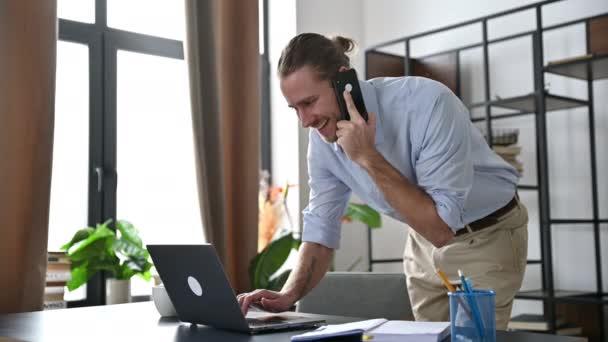 Satisfeito ocupado caucasiano bem sucedido empresário ou freelancer trabalhando em casa ou escritório, falando ao telefone com um colega enquanto usa um laptop, navegando na internet, respondendo a um e-mail — Vídeo de Stock