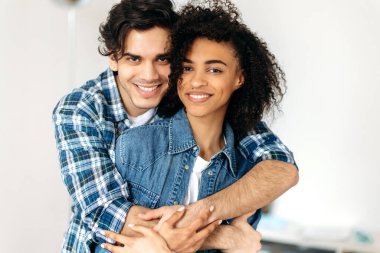 Birbirine aşık genç, çok ırklı bir çiftin portresi. Güzel Afro-Amerikan kıvırcık kızı ve Latin asıllı çekici adam birbirlerine sarılıyorlar, kameraya bakıp gülümsüyorlar, birlikte mutlular.