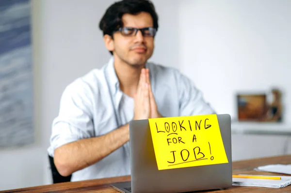 A clever guy, student or unemployed, sits at a table, looking for a job on the net, looks at advertisements, hopes to get a dream job, on a laptop a sign with the inscription looking for a job — Stock Photo, Image