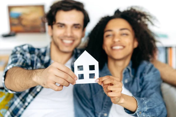 New home owners. Joyful multiethnic couple holding paper house, sitting on a sofa in their new modern apartment or house. Defocused african american girl and hispanic guy happy to buy their own home