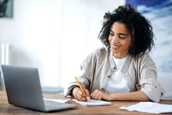 Enseignement à distance, leçon en ligne, travail à distance. Étudiante afro-américaine intelligente ciblée ou pigiste, dans un vêtement élégant, étudie ou travaille à distance, utilise un ordinateur portable, prend des notes lors d'une conférence en ligne — Photo