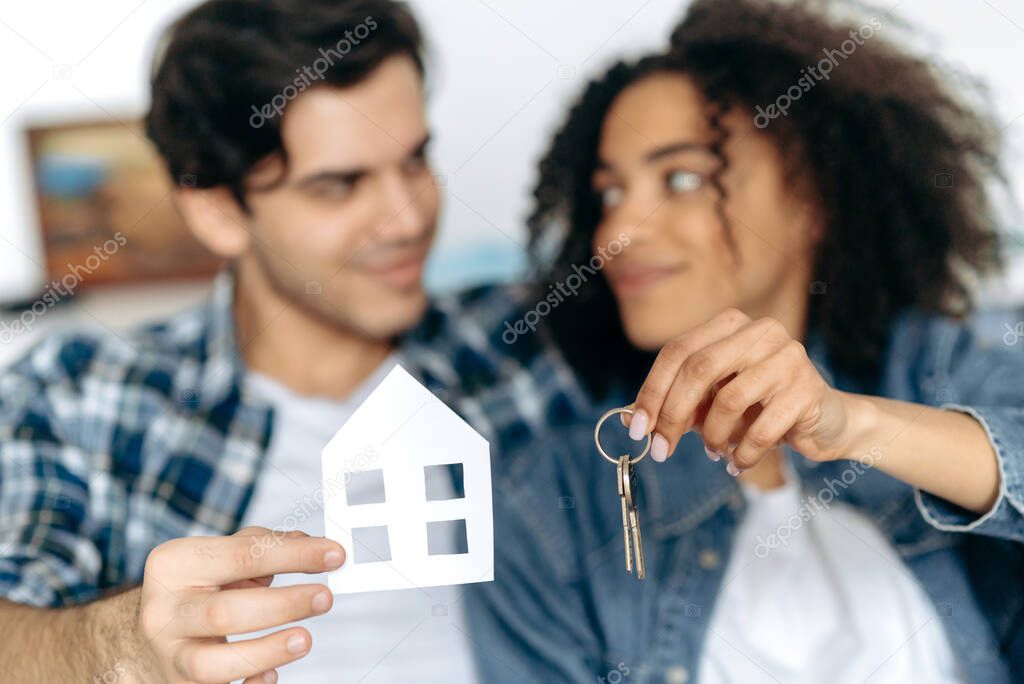 Purchase of new housing. Defocused loving multiracial happy couple became the owner of their first house, holding a small paper house and keys to new apartment, smiling, happy together