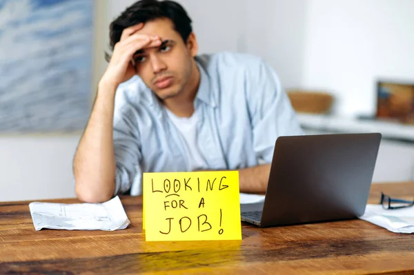 Ragazzo sconvolto disperato, studente, disoccupato, stanco di cercare un lavoro, seduto alla sua scrivania, guardando tristemente di lato, c'è un cartello sul tavolo con l'iscrizione in cerca di un lavoro. — Foto Stock