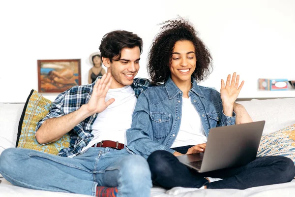 Feliz joven pareja multirracial pasa tiempo libre en casa en el sofá, utilizando un ordenador portátil, que habla a través de videoconferencia con la familia o amigos, interlocutor de saludo, sonriendo, felices juntos — Foto de Stock