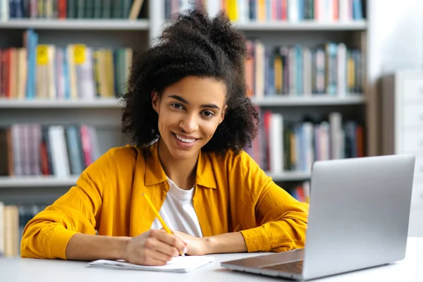 Retrato de menina adolescente afro-americana. Estudante de raça mista agradável sentado em uma mesa na biblioteca, com um laptop, tomando notas durante uma palestra, olhando para a câmera, sorrindo — Fotografia de Stock