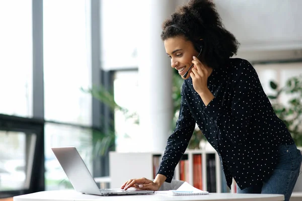 Occupé succès afro-américaine jeune femme d'affaires, gestionnaire ou consultant, dans un casque, se tient près d'un bureau dans un bureau, utilise un ordinateur portable, dactylographier un message, ou communiquer avec un client en ligne — Photo