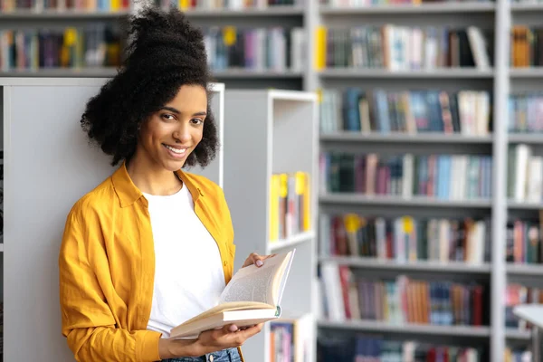 一个年轻美丽的非洲裔美国女人的画像时尚的女学生站在图书馆时，手里拿着一本书，准备上课或考试，看着相机，面带微笑 — 图库照片